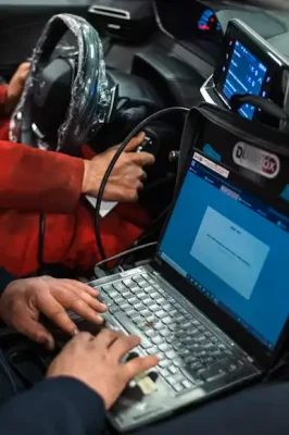Mobile mechanic running a car diagnostic scan with a laptop and diagnostic tool inside a vehicle with a protected steering wheel.
