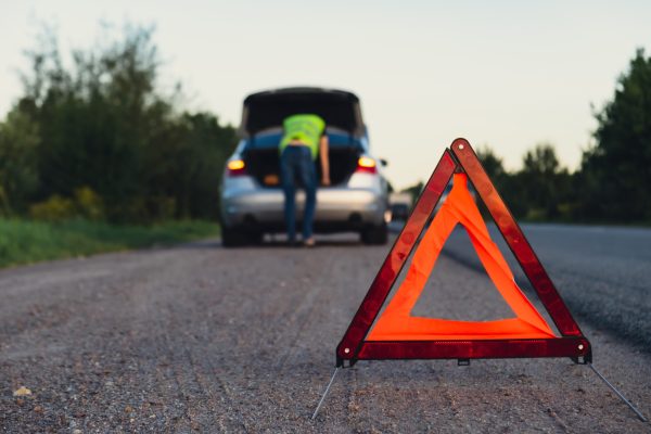 Unrecognizable sad driver in reflective vest. Male driver standing near broken car with open up hood. Red triangle to warn other road users of car breakdown or engine failure stop at countryside highway. Emergency traffic situation