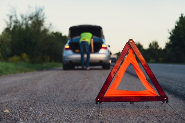 Unrecognizable sad driver in reflective vest. Male driver standing near broken car with open up hood. Red triangle to warn other road users of car breakdown or engine failure stop at countryside highway. Emergency traffic situation